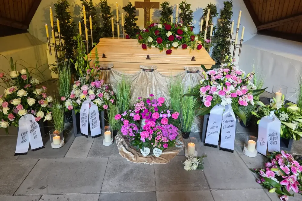 Heller Holzsarg mit rosa und weißen Blumen unter einem Holzdach, Trauerfeier von Bestattungen Schwarz