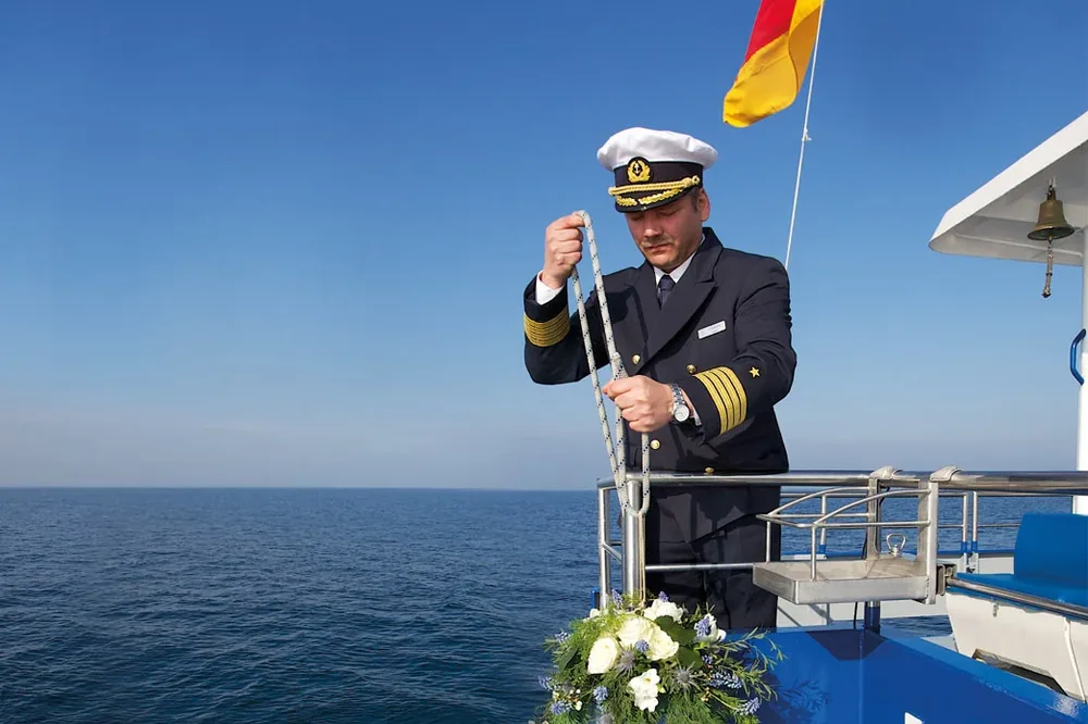 Kapitän an Bord eines Schiffes mit einer blauen, blumengeschmückten Urne, dahinter das offene Meer