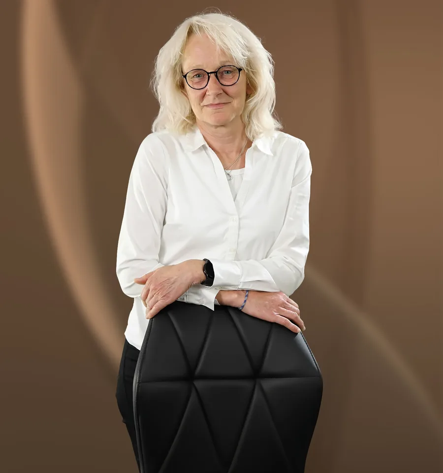 Angelika Roßmair, eine Frau in weißem Hemd und mit Brille, neben einem schwarzen Stuhl