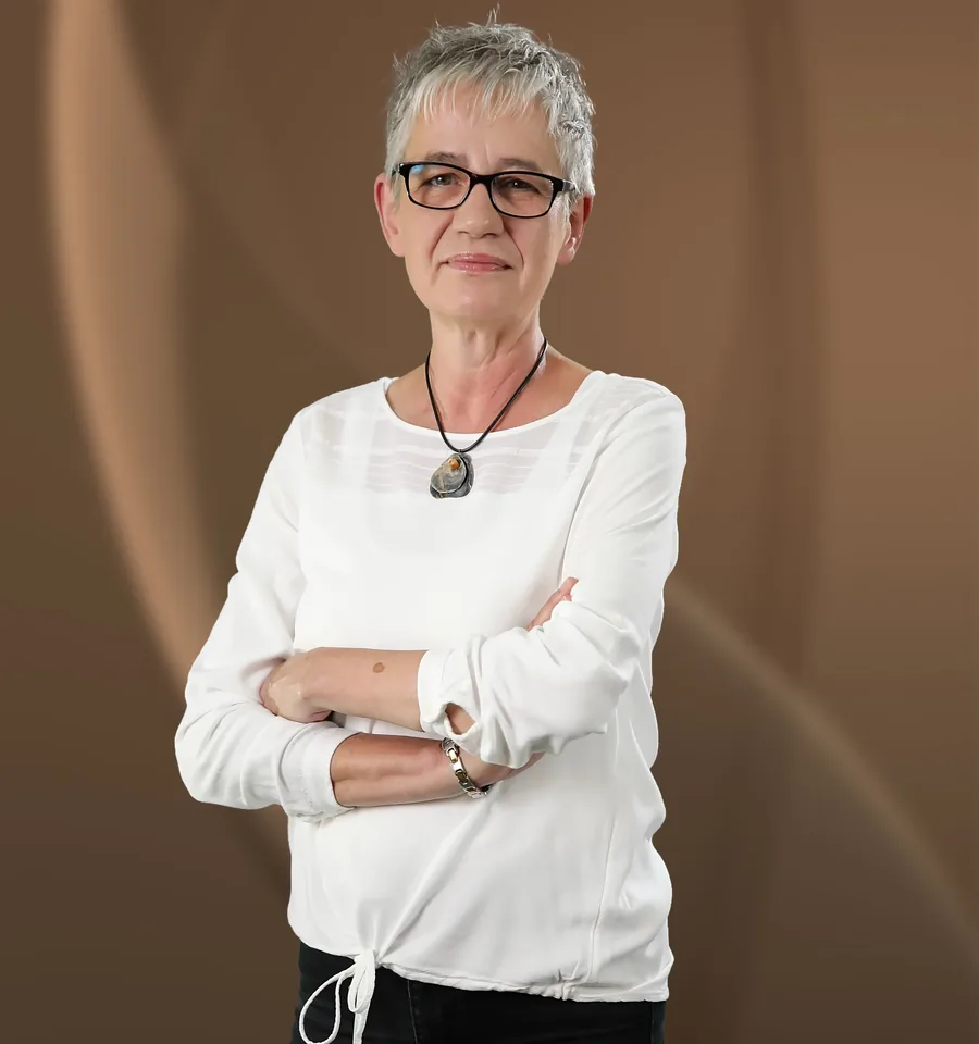 Rosi Müller, eine Frau mit Brille und weißem Hemd, vor braunem Hintergrund