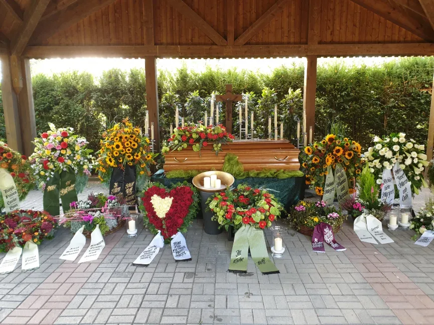 Holzsarg unter einem Holzpavillon, umgeben von Blumenkränzen und Kerzen