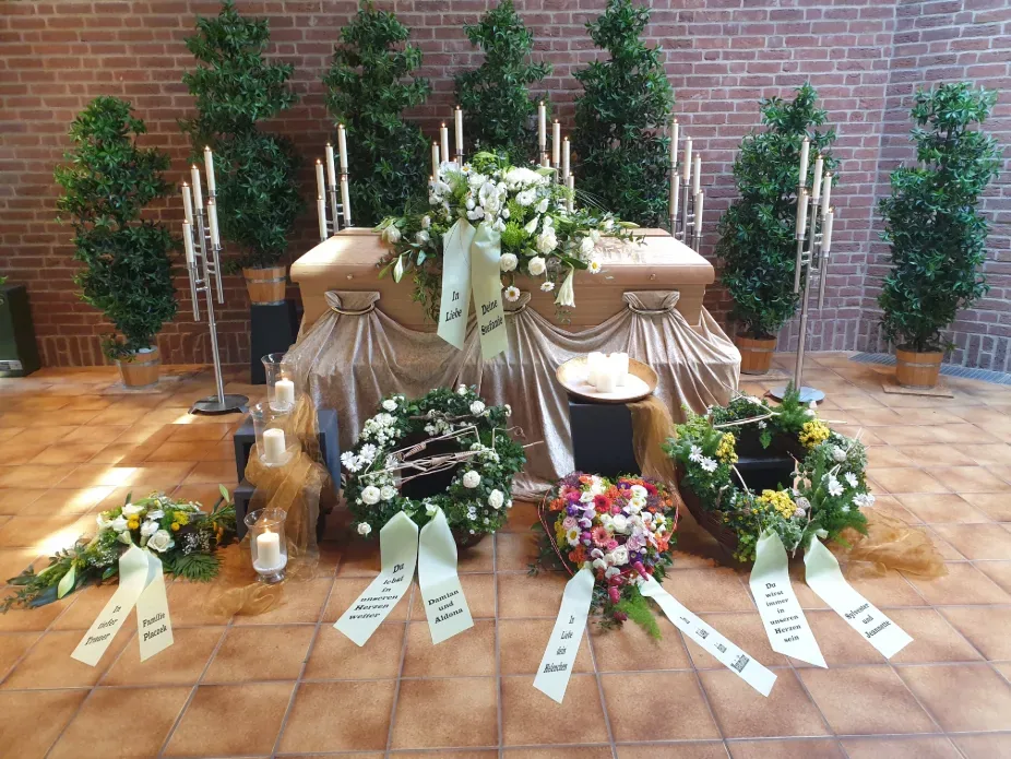 Trauerarrangement mit weißen Blumen, Kränzen und Kerzen vor einer Backsteinwand, gestaltet von Bestattungen Schwarz